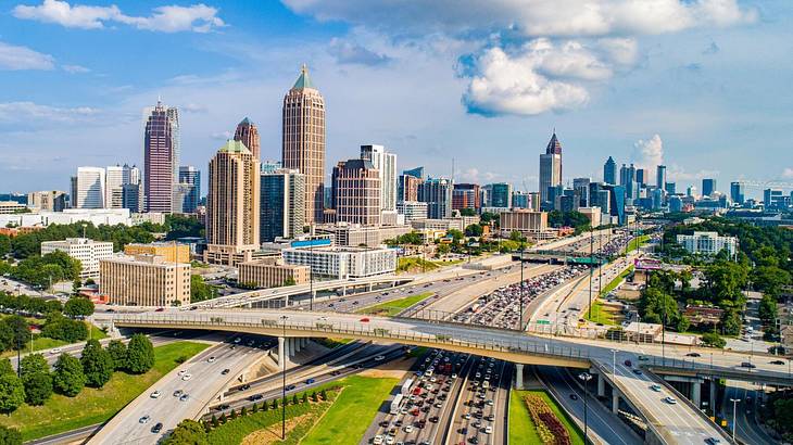 City buildings next to trees, grass, and a highway under a blue sky with clouds