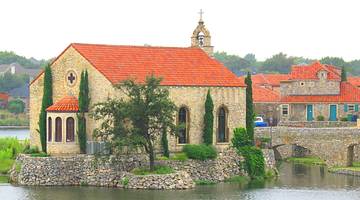 A European-style stone building with an orange roof next to water and green plants
