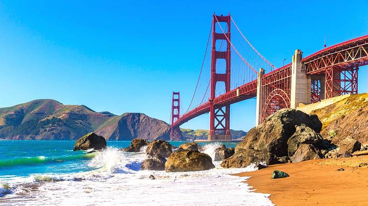 A red suspension bridge over a bay with a small beach with sand and rocks in front