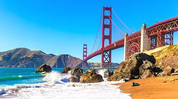 A red suspension bridge over a bay with a small beach with sand and rocks in front