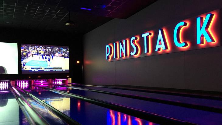An illuminated bowling alley with a neon sign that says "Pinstack" on the wall