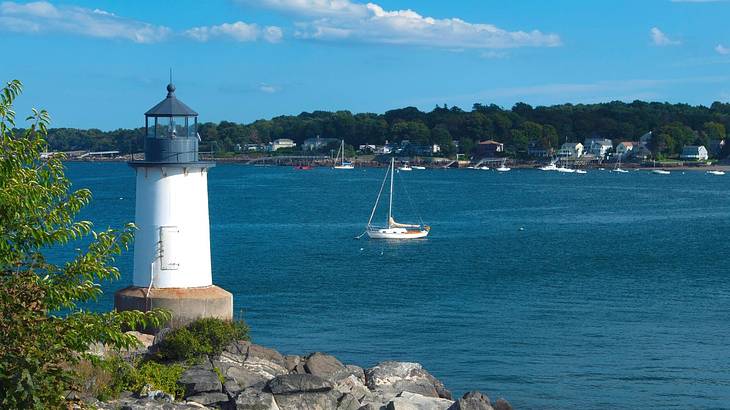 A small white lighthouse next to a bay with a boat on the water and greenery opposite