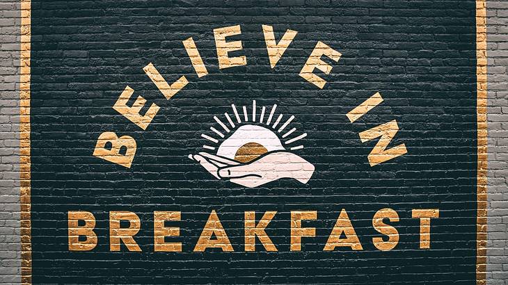 A black sign with gold trim painted on a brick wall that says "Believe in Breakfast"
