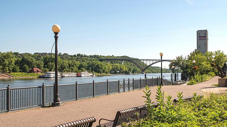 A walkway along a river with greenery surrounding it and a bridge in the distance