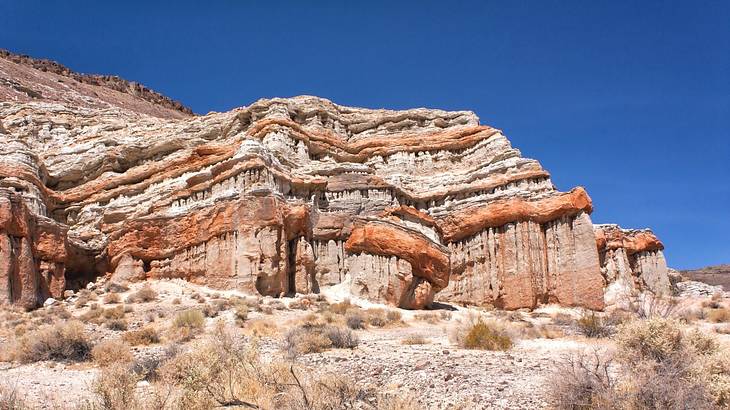 A red rock mountain with sand and shrubs in front of it under a blue sky