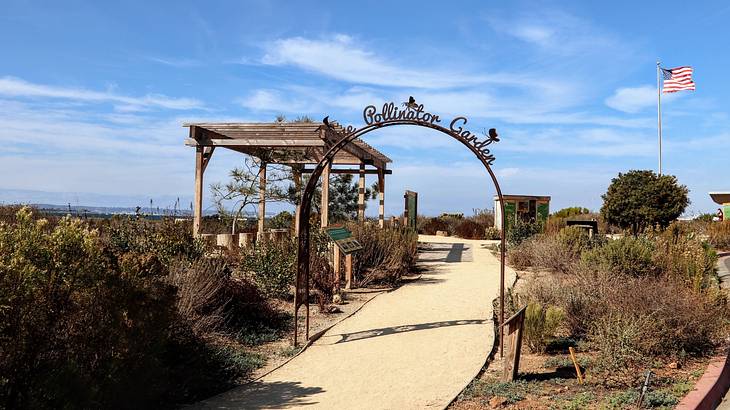 A path through a garden with an arched gate that says "pollinator garden"