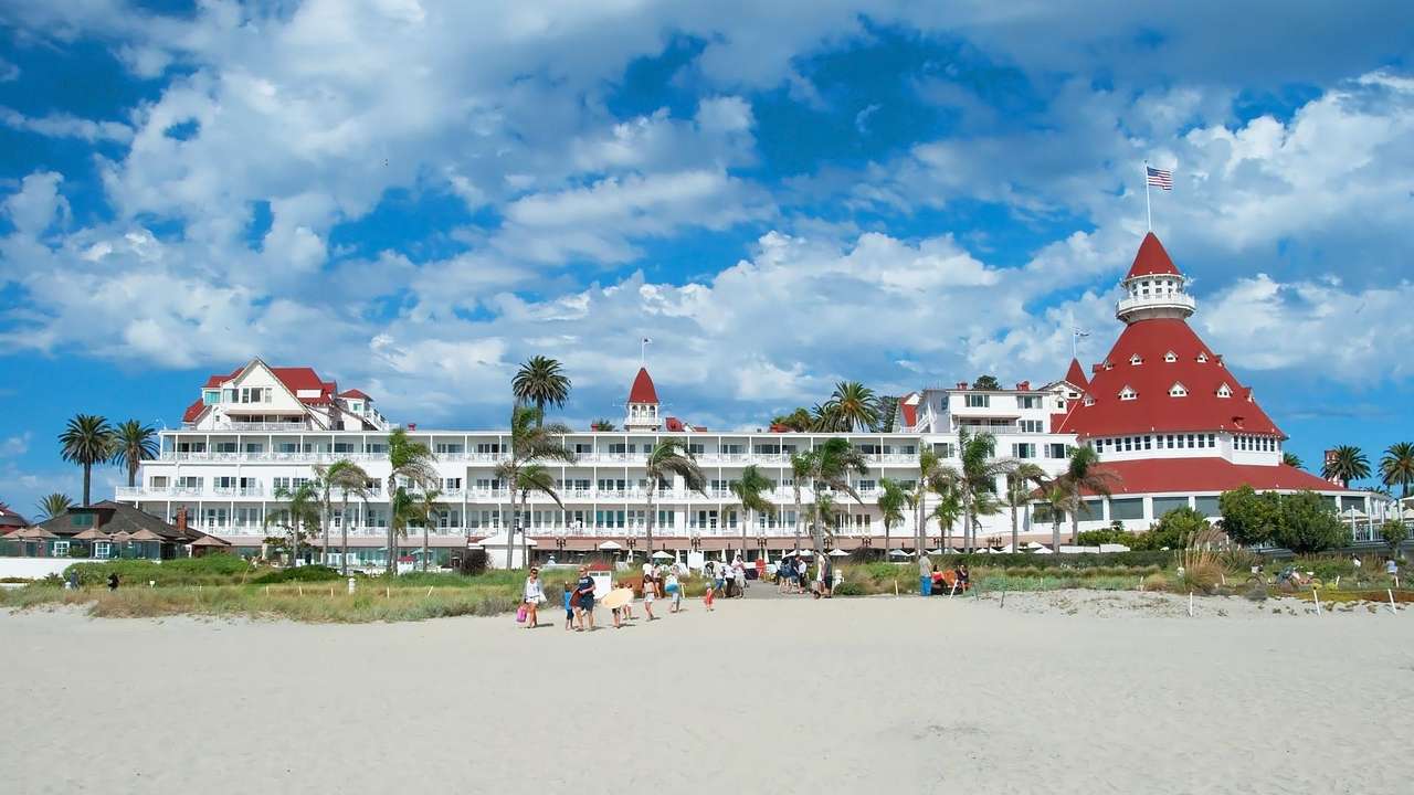 A white hotel with a large red domed roof next to a sandy beach and palm trees