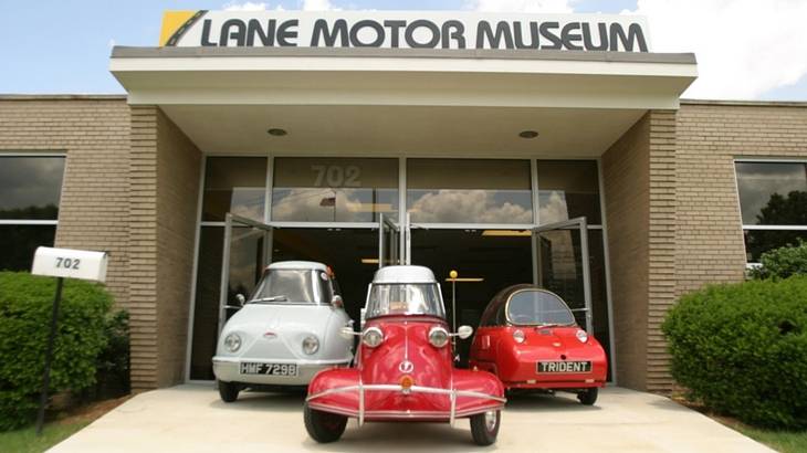 Three antique cars outside of a building with a "Lane Motor Museum" sign