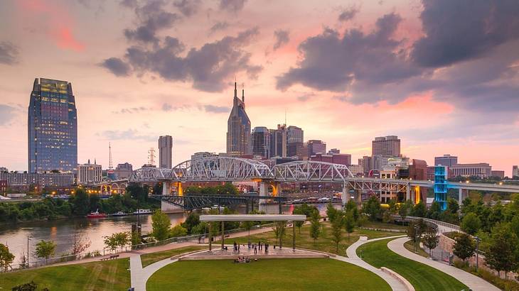 A city skyline and a bridge over a river next to a green park under a pink sky