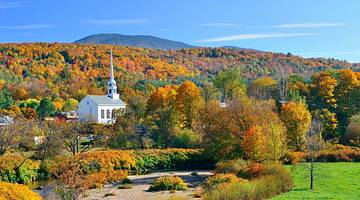 A white church surrounded by orange fall trees and green grass on a clear day