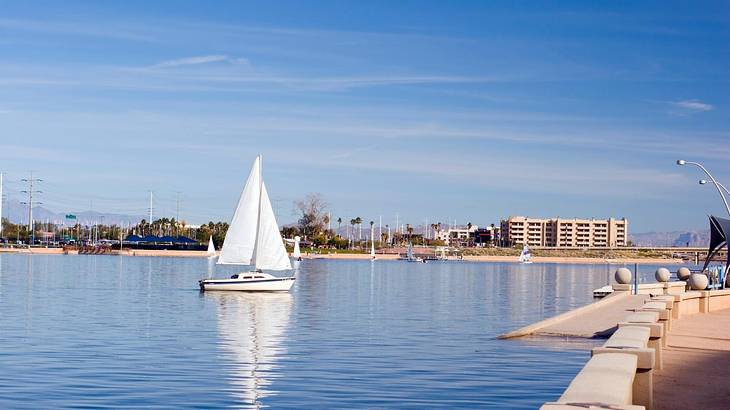 A sailboat on the water with buildings in the distance and a path to the side A sailboat on the water with buildings in the distance and a path to the side