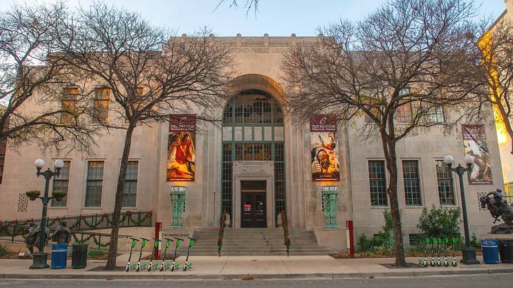 A stone museum building with banners on it and bare trees on the street in front