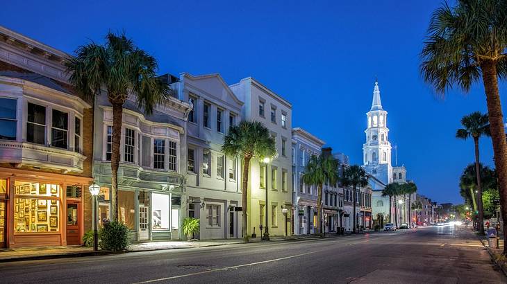 A city street at night with palm trees, illuminated buildings, and a church