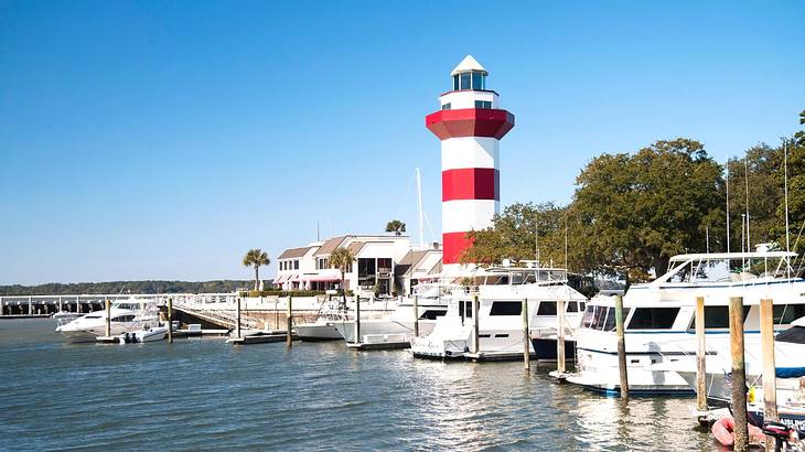 A marina with boats docked next to a white and red striped lighthouse and trees