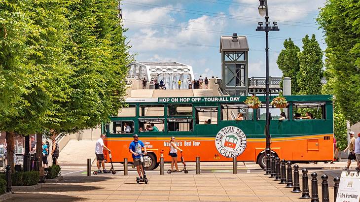 A green and orange sightseeing bus with a path, trees, and people around it