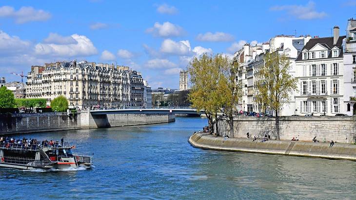 A river with a boat on it and trees and white Parisian buildings on the riverside