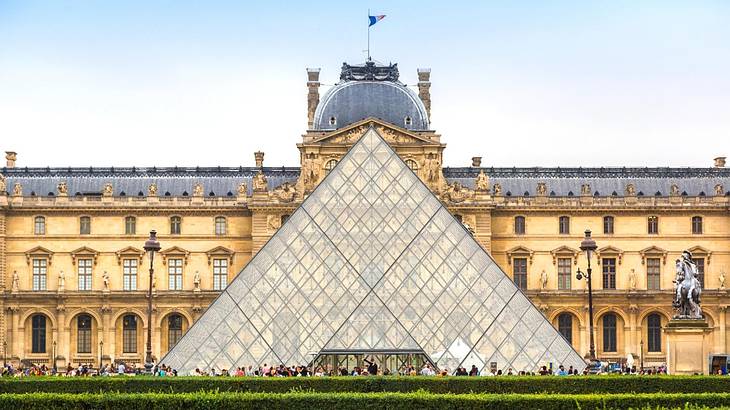 A glass pyramid structure with a stone mansion behind it and a green hedge in front