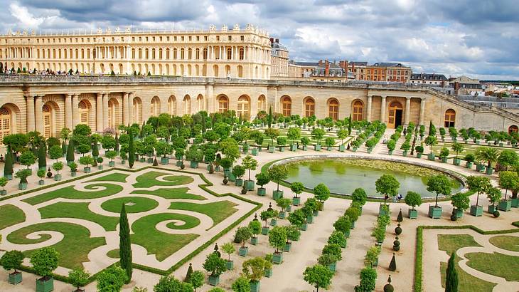 A manicured garden with mazes, trees, and a pond, and a stone building behind it