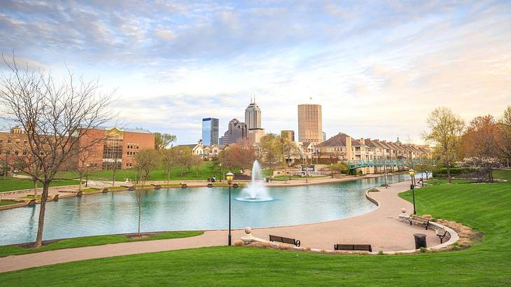 A pond with a fountain and grass surrounding it and a skyline in the distance