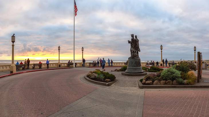 A beachside promenade with a flag, lamppost, and statue on it at sunset