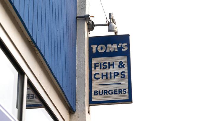 A blue sign on a building with blue paneling that says "Tom's Fish & Chips"