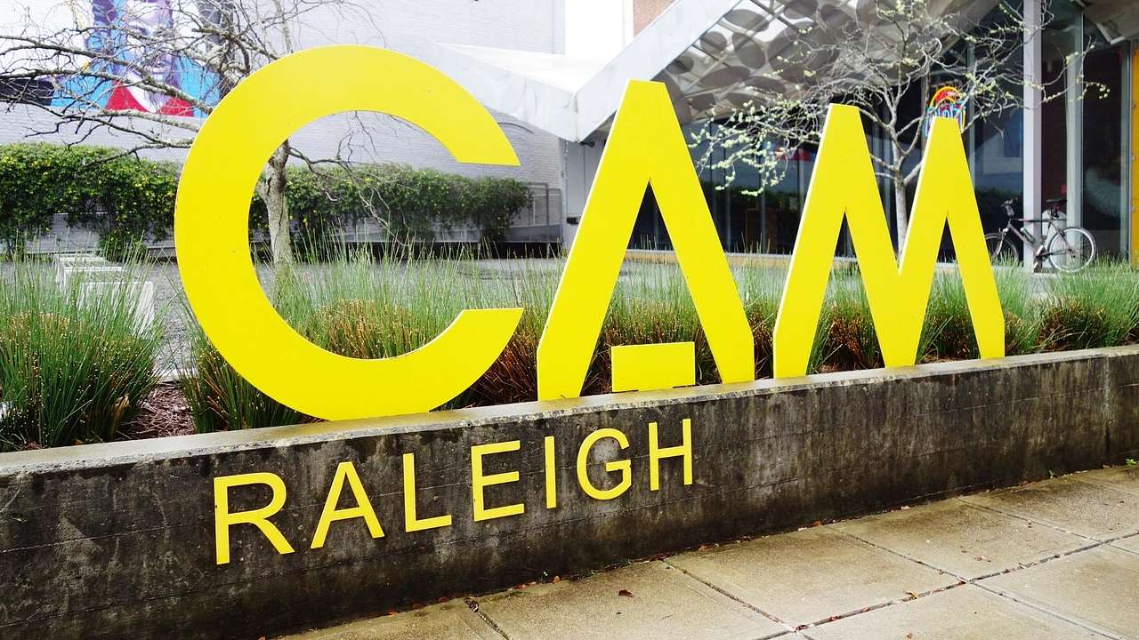 A yellow sign that says "CAM Raleigh" on a wall with a garden behind it