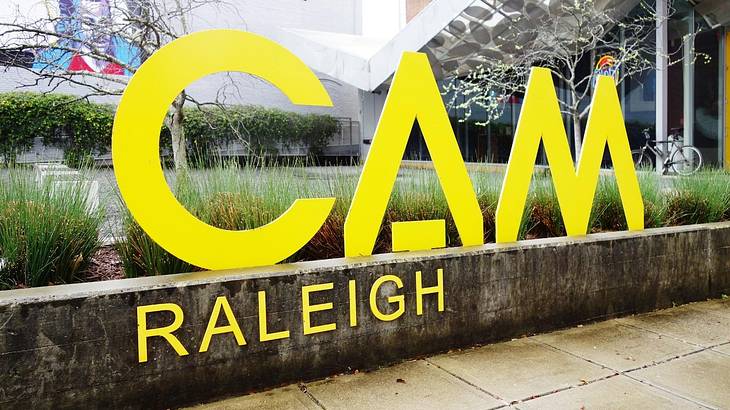 A yellow sign that says "CAM Raleigh" on a wall with a garden behind it