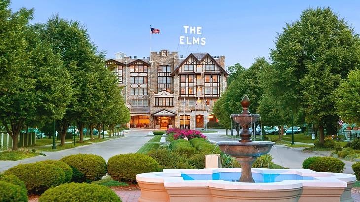 A hotel with a "The Elms" sign and a path, trees, and water fountain in front of it