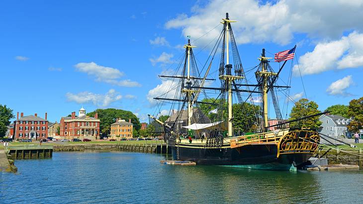 An old-fashioned ship on the water with buildings and grass on the shore