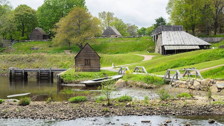 Old wooden buildings on grass with a river in front of them and trees surrounding