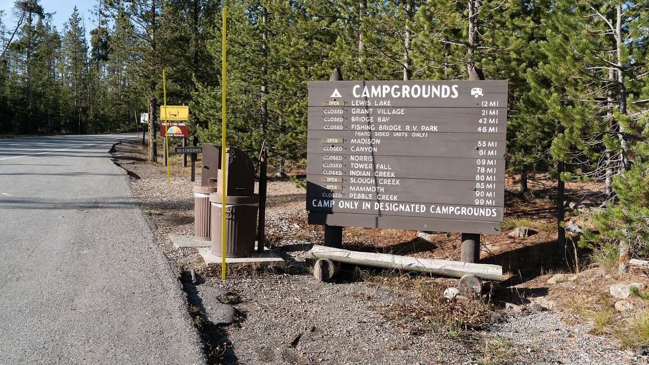 Tree-lined dirt road and brown campground sign with many sites written in white
