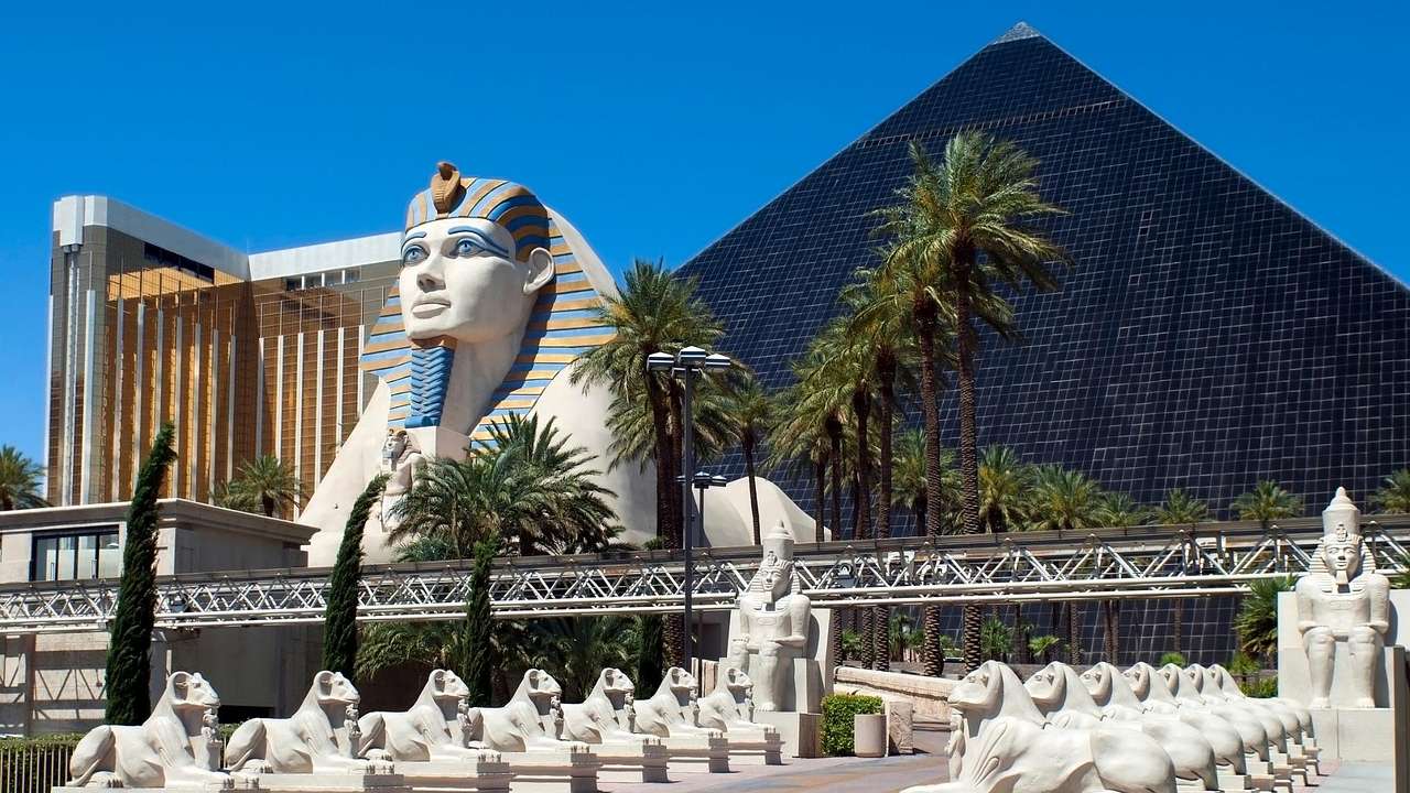 A Sphinx statue, a dark glass pyramid replica, with palm trees surrounding them