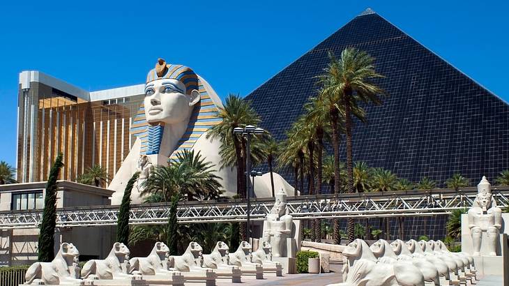 A Sphinx statue, a dark glass pyramid replica, with palm trees surrounding them