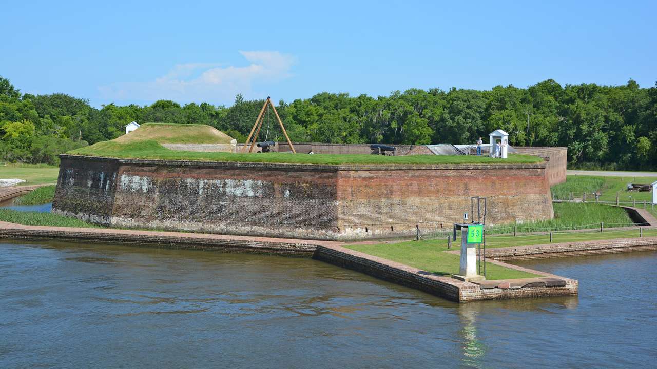 A small old fort with greenery on top and surrounding and water in front