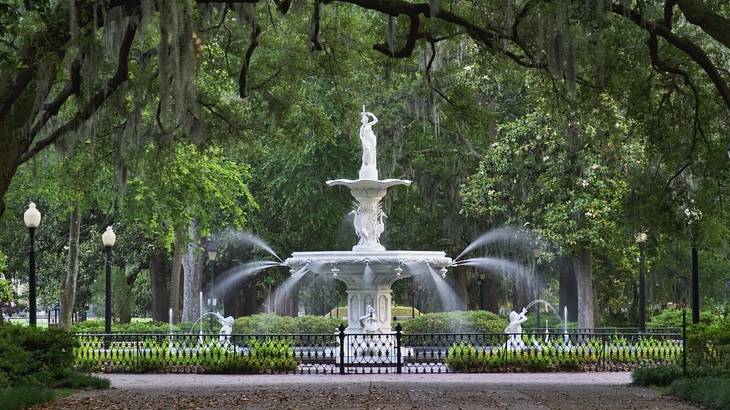 A water fountain with a path and iron gate in front surrounded by trees