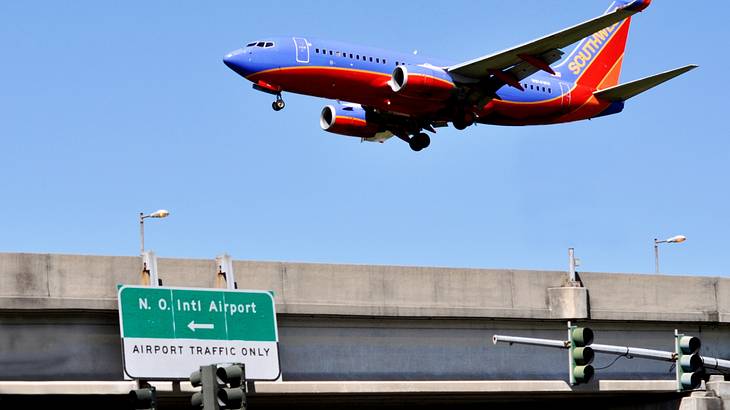 A blue and red plane flying next to an overpass with a green "N.O. Intl Airport" sign