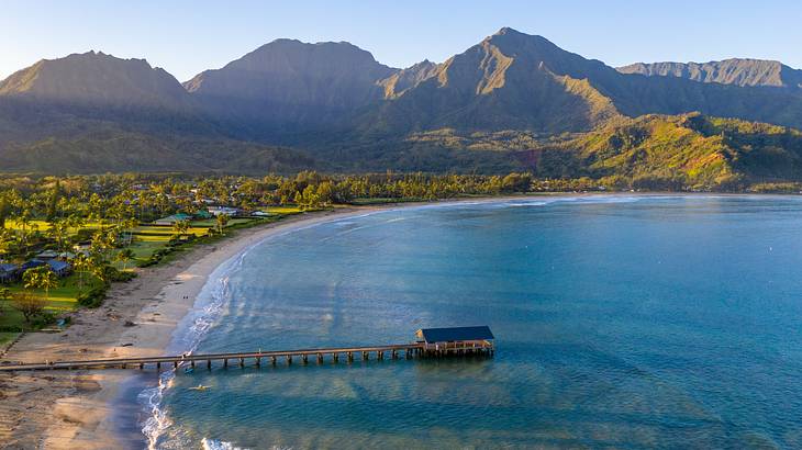 A beach with a small hut in the water and greenery mountains surrounding