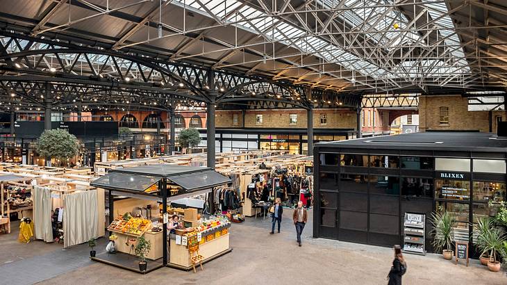 Inside a market with a fruit kiosk and lit-up stores under an industrial roof