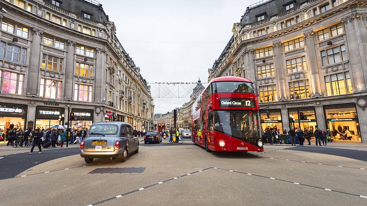 A street with old buildings, lit-up shops, a double-decker bus, & cars