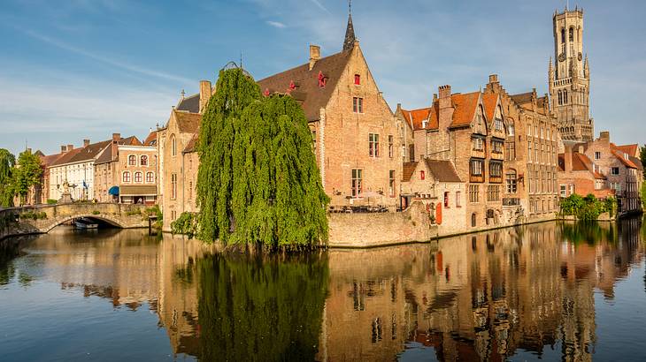 Medieval buildings, greenery, and a stone bridge on the left reflected in a canal