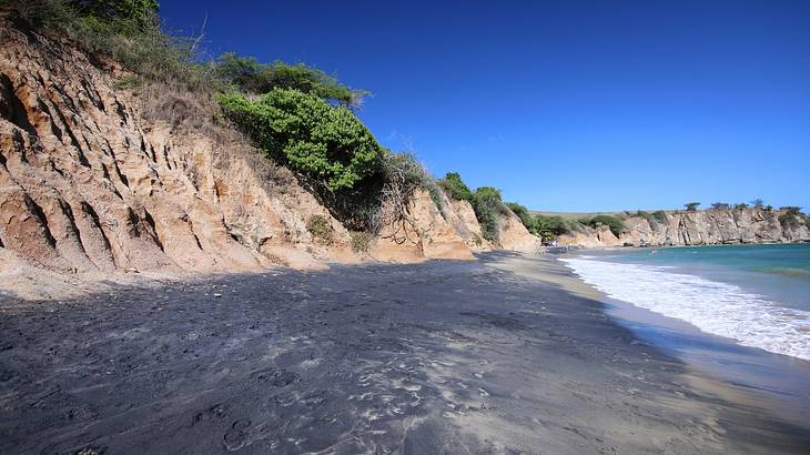 Cliff with vegetation along a sandy black beach, under a vibrant blue sky