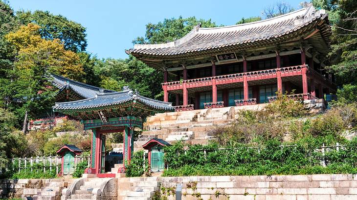 A small red temple gate below a bigger temple with steps, surrounded by greenery