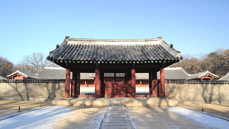 A small red temple with a black roof next to bare winter trees and a cement wall