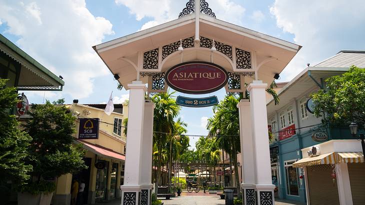 An arch with a sign that says Asiatique in front of trees and shops
