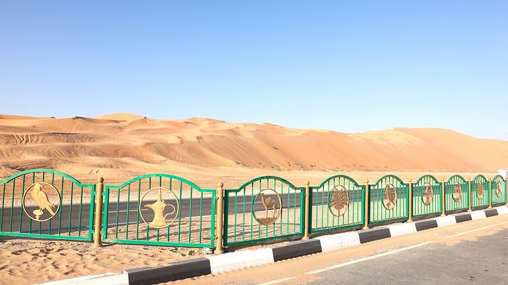 Sand dunes behind a turquoise and gold fence on a clear blue day