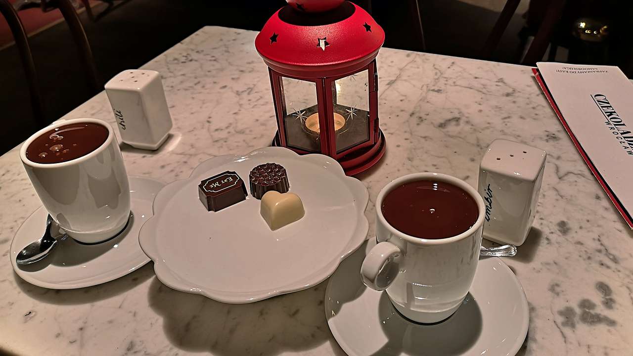 A marble table with two mugs of hot chocolate, chocolates and a red lantern
