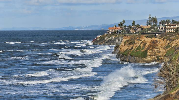 Waves crashing against rocky cliffs with ocean-view mansions at the top