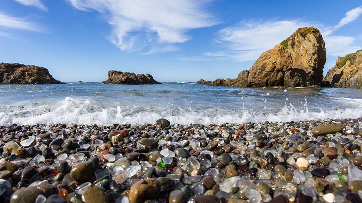 Breaking waves on a beach covered in multicolored sea glass and rocky cliffs behind