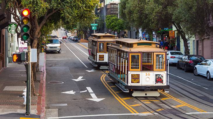 Two cable cars in the middle of a street with trees and parked cars