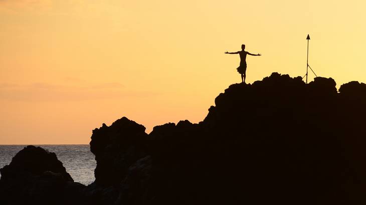 Rocks in the dark with a person standing on top in front of an orange sunset